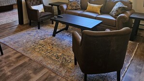 Un salon avec un canapé en cuir marron, un fauteuil marron foncé et une table basse. Une grande fenêtre offre une vue sur la verdure à l’extérieur.