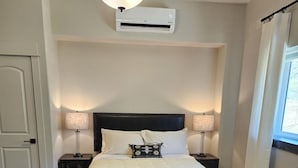 A bedroom with a bed, two bedside tables with lamps, an air conditioning unit, a door, and a window with curtains. - Your Ideal Retreat! Onsite Parking, Near Gardiner Downtown! (Gardiner)