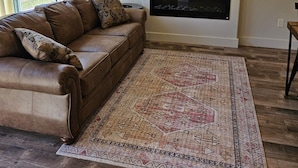 A living room with a brown leather sofa, a patterned rug, a flat-screen TV mounted above a fireplace, and a window with curtains. - Your Ideal Retreat! Onsite Parking, Near Gardiner Downtown! (Gardiner)