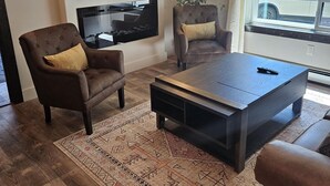 A modern living room with a large flat-screen TV mounted on the wall, a dark coffee table, two leather armchairs with cushions, and a patterned rug on the wooden floor.