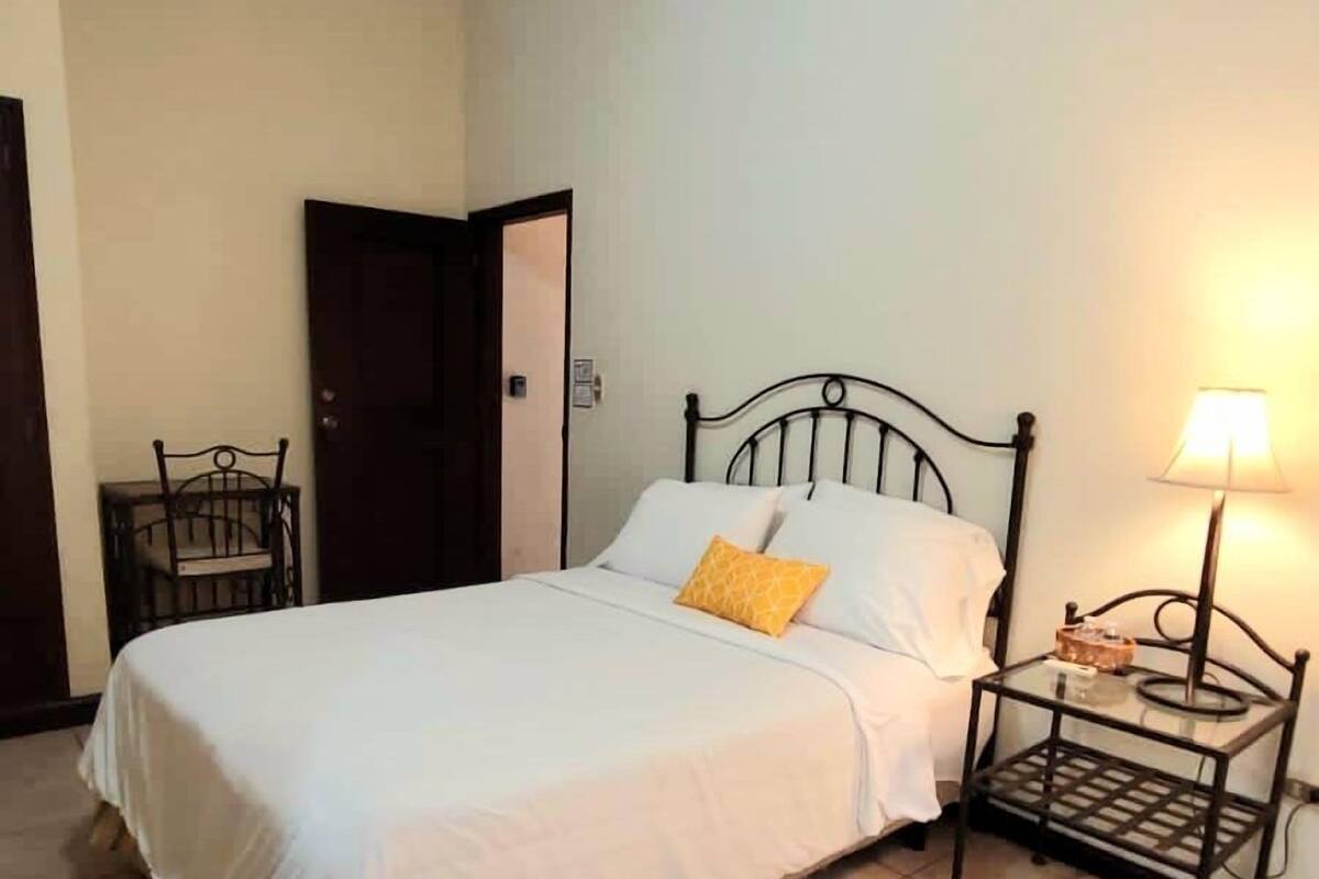 A single bed with a white bedspread and a yellow pillow, a small bedside table with a lamp, and a chair in a room with tiled flooring and a door in the background.