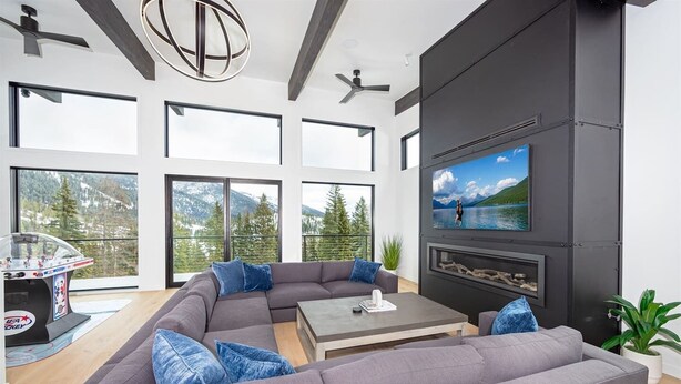 A modern living room with a sectional sofa, a coffee table, and a fireplace. Large windows offer a view of mountains and trees.