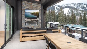 A modern outdoor living area with a stone fireplace, a large flat-screen TV, and a wooden bench.