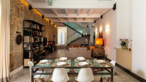 A dining area with a long table set for four, flanked by white chairs. A staircase is visible in the background leading to an upper level. - Valverde Art Space by DomuSicily (Palermo)