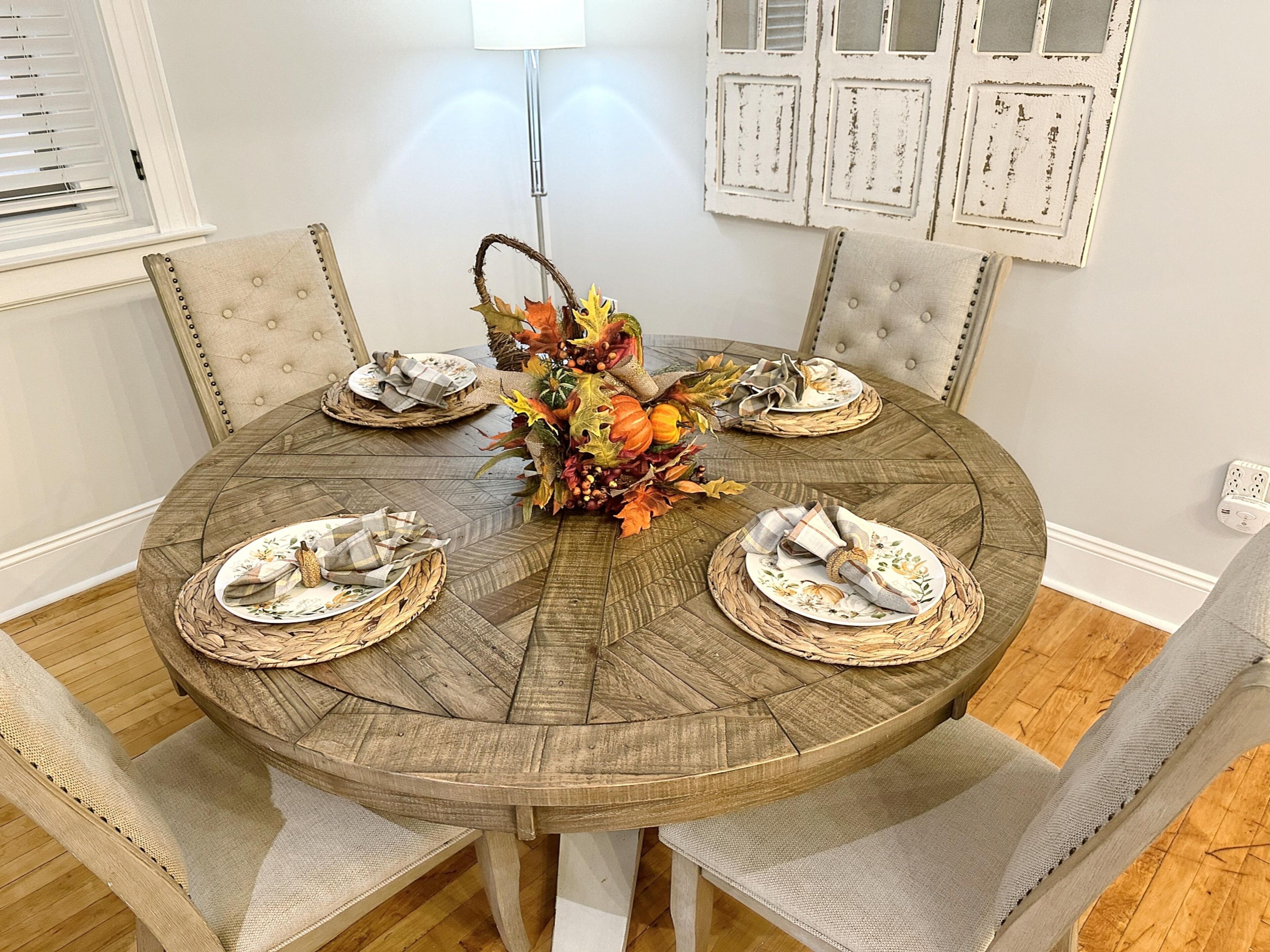 Une table de salle à manger ronde en bois, accompagnée de quatre chaises, d’un set de table en fibres naturelles et d’un centre de table décoré de feuilles d’automne.