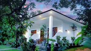 A white building with a covered porch, surrounded by lush greenery and potted plants. - The Cattleya Guest House  (Sigiriya)