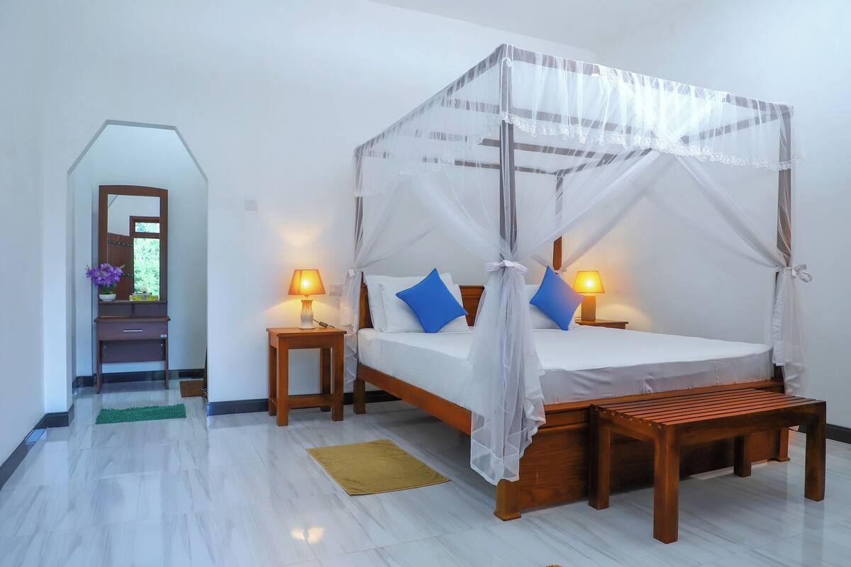 A bedroom with a four-poster canopy bed, two bedside tables with lamps, a wooden bench, and a green mat on the floor.