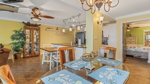 A dining area with a glass table, chairs, and a chandelier. There is a kitchen in the background with a refrigerator and cabinets.