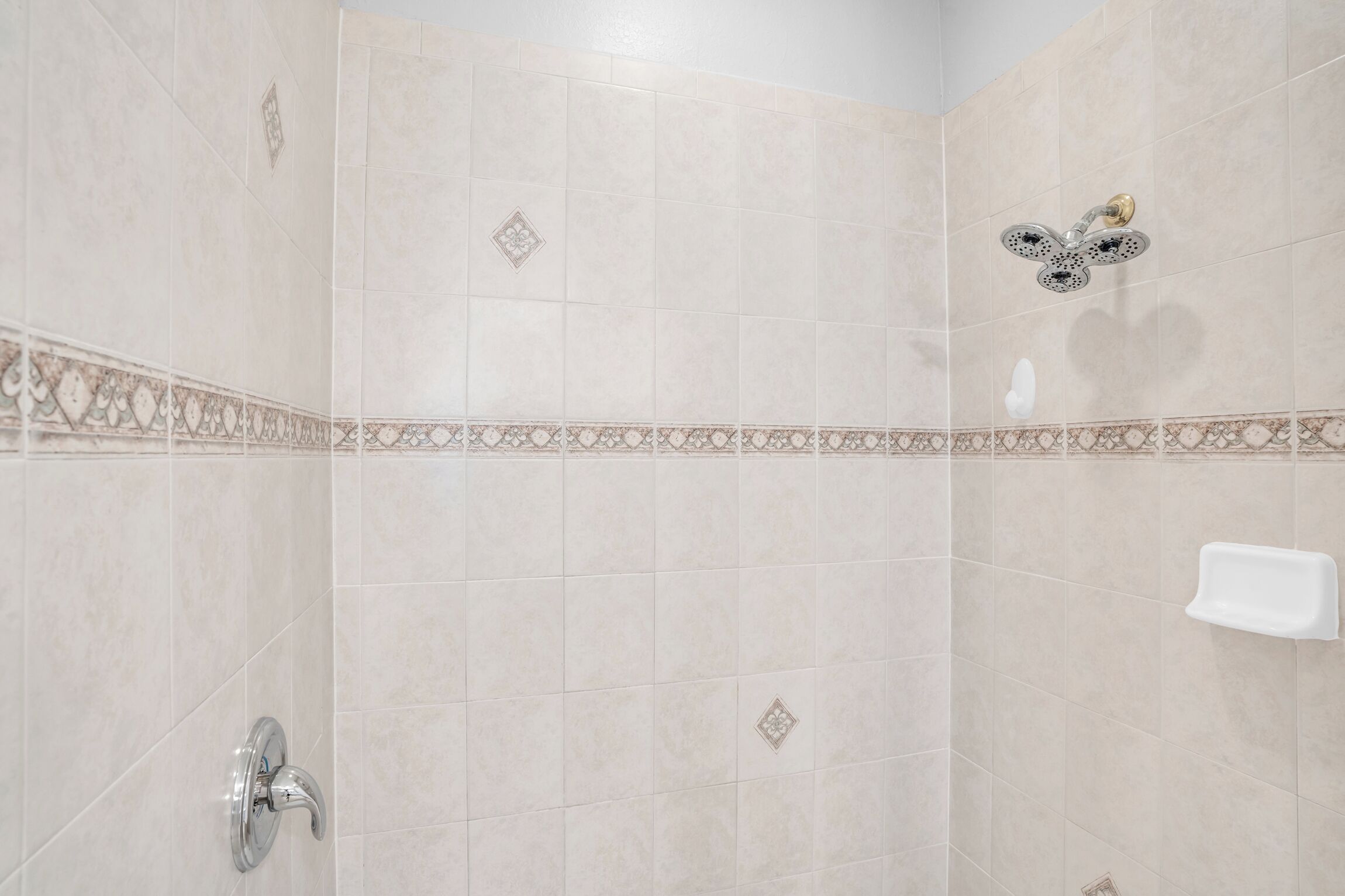 A bathroom with beige tiles, a shower with a handheld showerhead, and a soap dish on the wall.