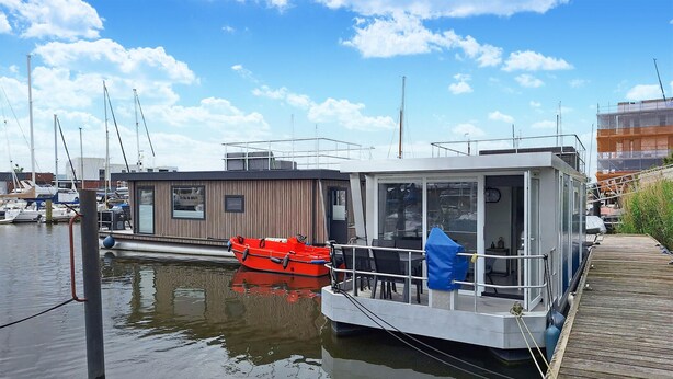 Een drijvend woonschip afgemeerd in een jachthaven, met een kleine rode boot in de buurt.