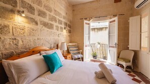 Un dormitorio con pared de piedra, una cama con ropa de cama blanca, una silla a rayas y un balcón con barandilla.