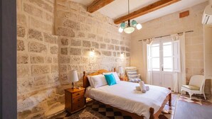 Un dormitorio con pared de piedra, cama de madera, mesita de noche, una silla y ventana con cortinas.