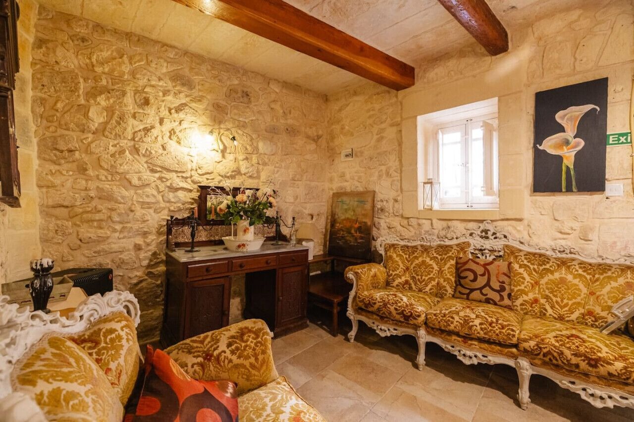 A stone-walled room with a wooden ceiling, a sofa, a small table with a vase, and a window with an exit sign.