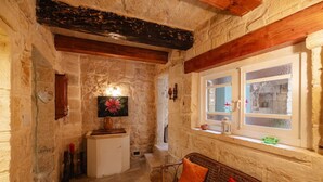 Una habitación rústica con paredes de piedra, techo de madera, un pequeño mueble blanco, un sofá de mimbre con cojines rojos y una ventana con vistas al exterior.