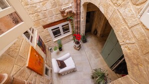 Un patio interior con muros de piedra, un banco acolchado blanco, plantas en macetas y una ventana con contraventanas.