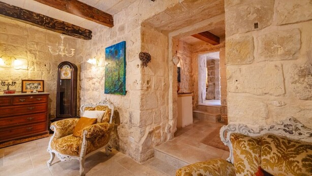 Una habitación rústica con paredes de piedra, techo de madera y un candelabro. Hay un sillón antiguo, un aparador de madera y un cuadro en la pared.