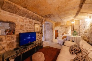 A stone-walled room with a television, a sofa, and a small table with a lamp. - Tranquil Haven suite in imdina (Ir-Rabat)