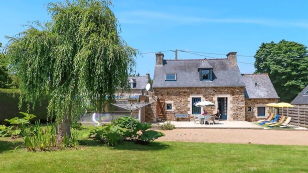 Une maison en pierre avec un grand arbre devant, une piscine et un espace salon extérieur.