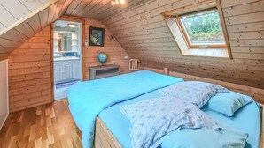Une chambre mansardée chaleureuse avec un plafond en pente en bois, un puits de lumière, un lit avec une literie bleue, un petit bureau avec un globe terrestre et une armoire blanche.