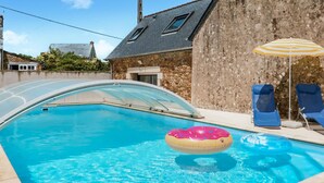 Une piscine recouverte d’un couvercle transparent, un flotteur rose gonflable et deux chaises longues bleues placées sous un parasol à rayures.