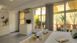 Une chambre d’hôtel moderne comprenant un lit, un bureau avec une télévision, un coin repas où le petit-déjeuner est servi, et un balcon agrémenté de plantes en pot.