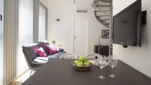 Un salon moderne avec un canapé gris, une table à manger agrémentée d’un panier de fruits et de verres à vin, un téléviseur fixé au mur et un escalier en colimaçon.
