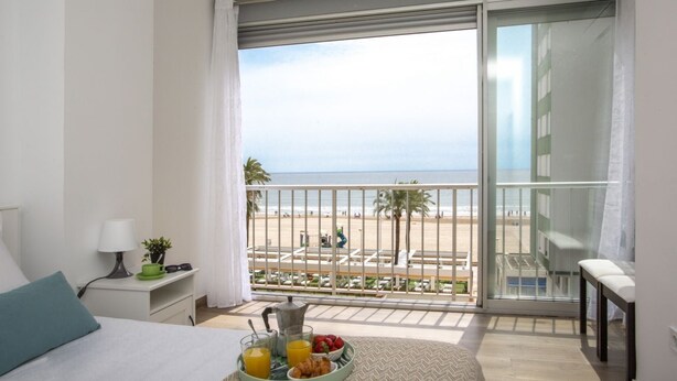 A balcony with a view of the beach, a breakfast tray with fruits and juice, and a bed with a pillow and a nightstand with a lamp and a plant.