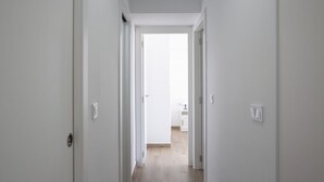 A narrow hallway with white walls and a wooden floor leading to a bright room with a door.