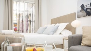 A hotel room with a bed, breakfast table with fruit and pastries, and a view of a street through the window.