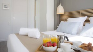 Un lit parfaitement fait, avec du linge de lit blanc, un plateau de petit-déjeuner comprenant des fraises, du jus d’orange et des croissants, ainsi qu’une table de chevet avec des tasses et une cafetière.