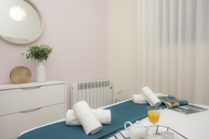 A hotel room with a bed, a bedside table with a vase of green plants, a radiator, a window with white curtains, and a breakfast tray with a glass of orange juice. - SingularStays Seu I (València)