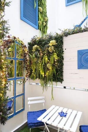 A cozy outdoor seating area with a white table and blue chairs, surrounded by lush greenery and decorative plants.