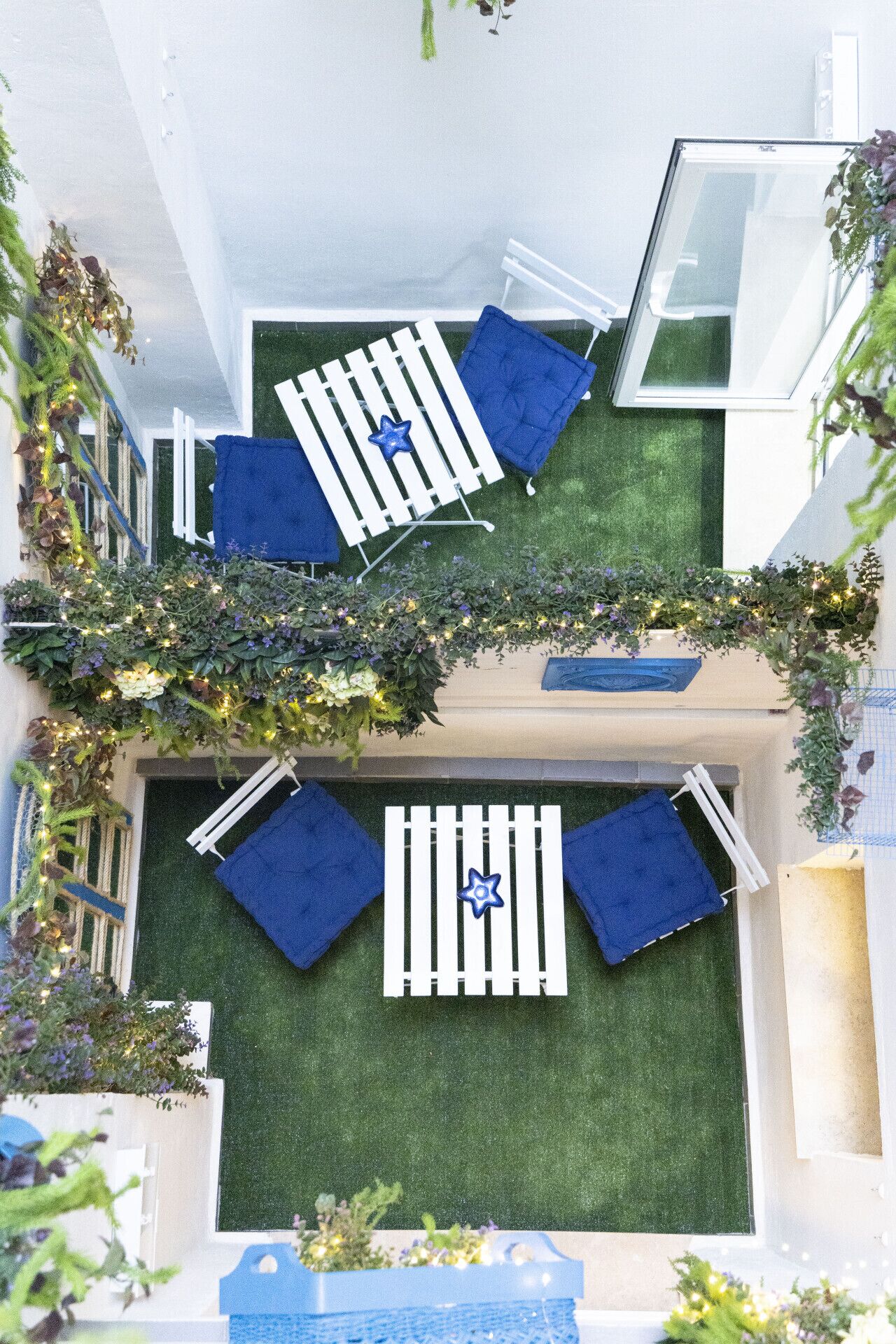 A rooftop garden with a small seating area, including a table and chairs, surrounded by greenery and decorative lights.