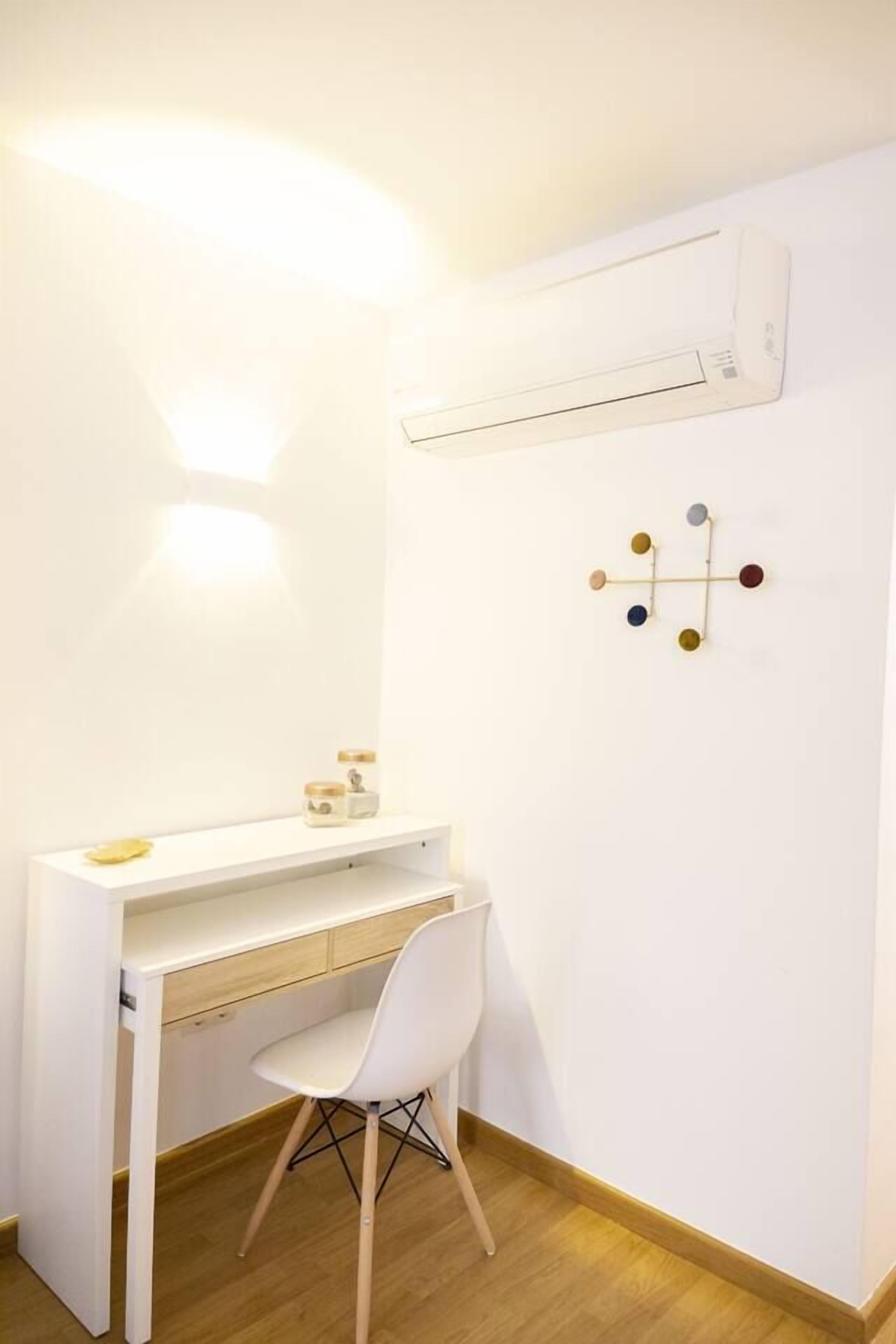 A compact room with a white desk, a white chair, a small shelf with items, a wall-mounted air conditioner, and a wall decoration.