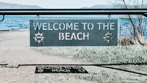 A welcome sign to the beach with a view of the sea and mountains in the background. - Water's Edge Suite 201 - Beautiful, Spacious & Luxurious (Garden City)