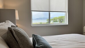 A bedroom with a bed, pillows, a nightstand, a lamp, and a window with a view of a lake and trees. - Water's Edge Suite 201 - Beautiful, Spacious & Luxurious (Garden City)