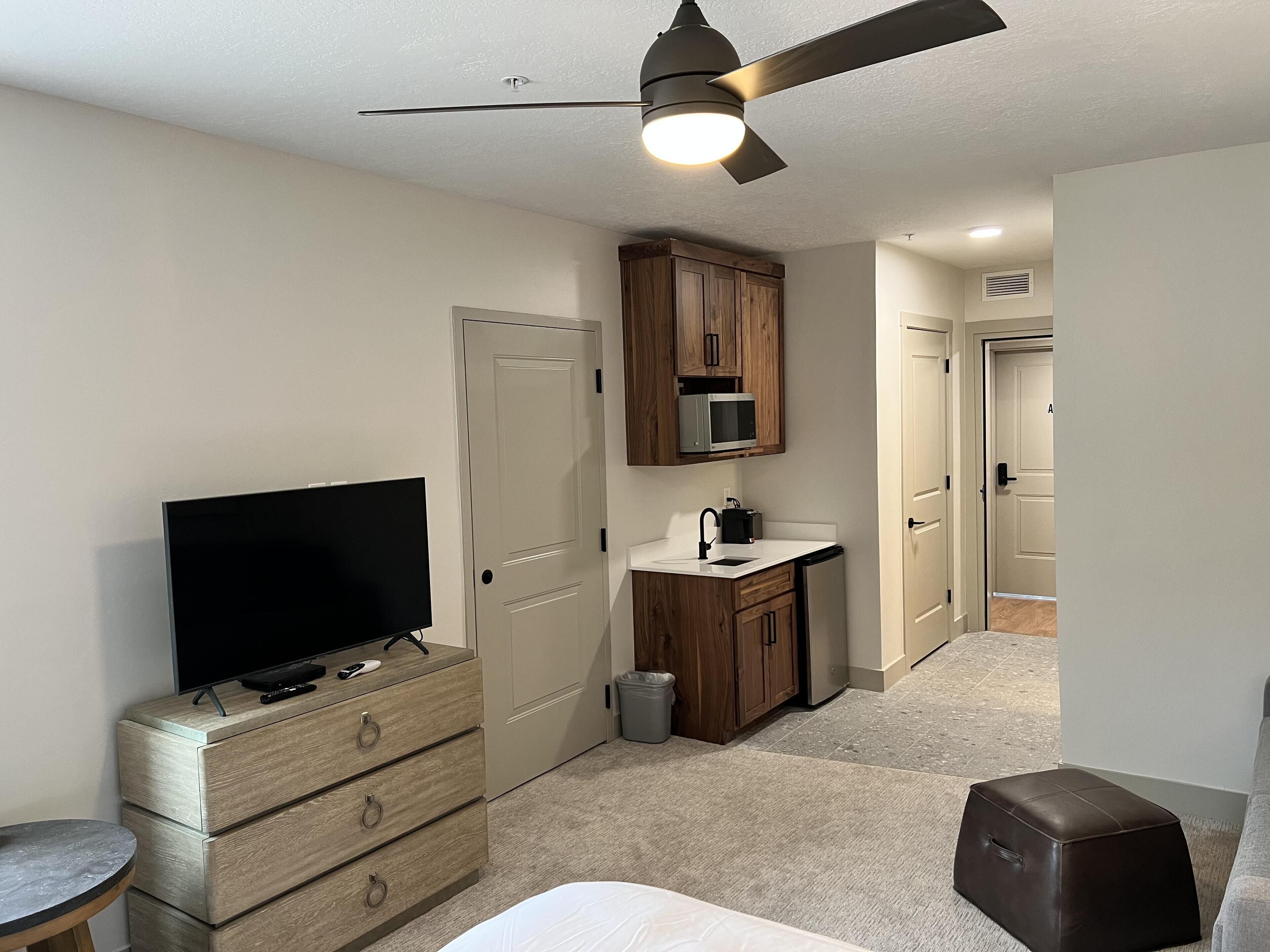 Habitación compacta con televisión de pantalla plana, armarios de madera, microondas, un lavamanos pequeño y un ventilador de techo.