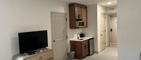 Habitación compacta con televisión de pantalla plana, armarios de madera, microondas, un lavamanos pequeño y un ventilador de techo.