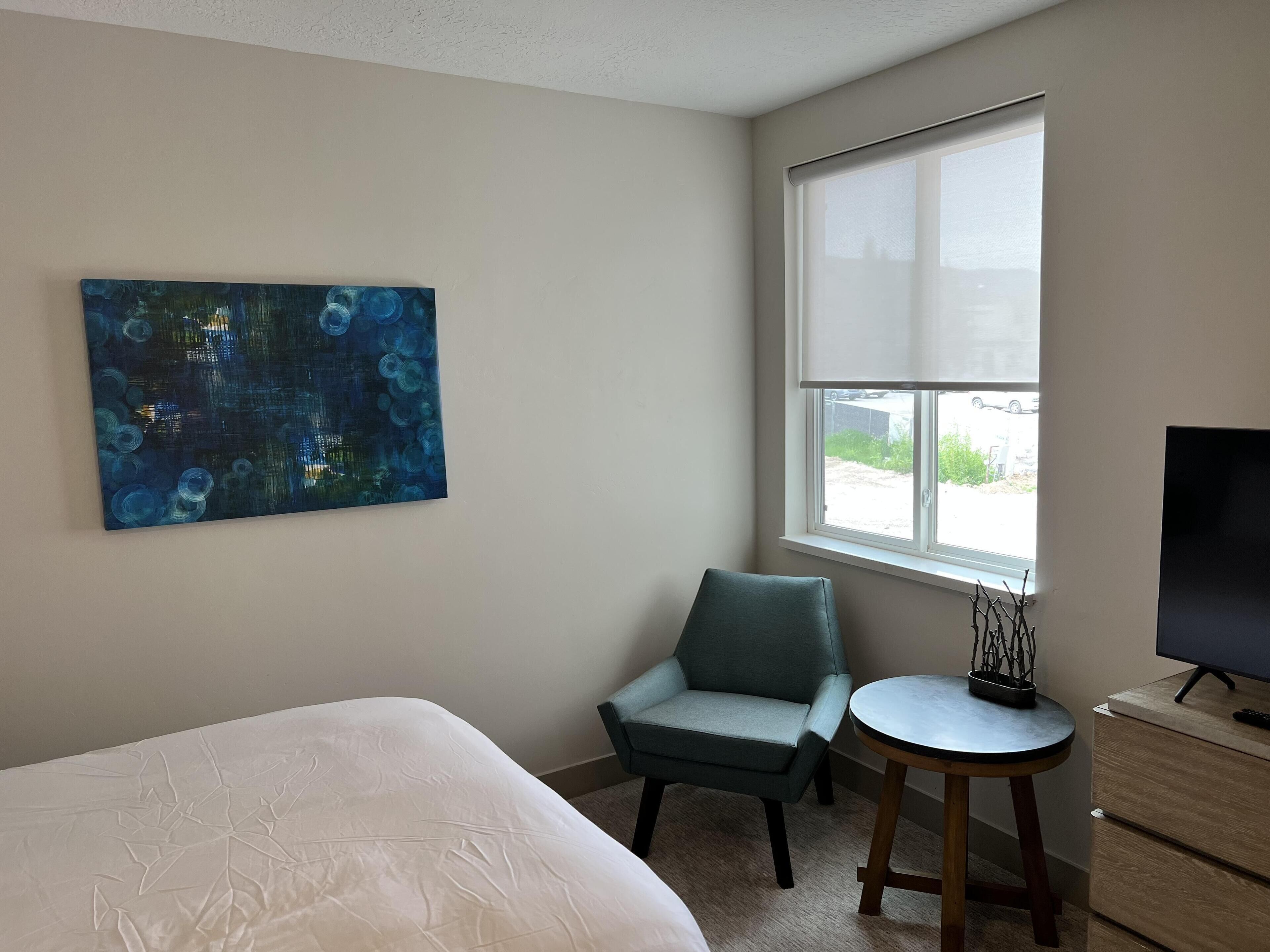 A bedroom with a bed, a chair, a small table, a TV, and a window with a view of the outside.