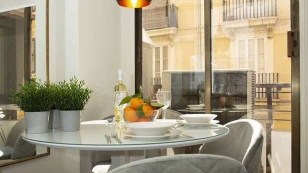 A dining area with a glass table, two potted plants, a bowl of fruit, and a bottle of wine.
