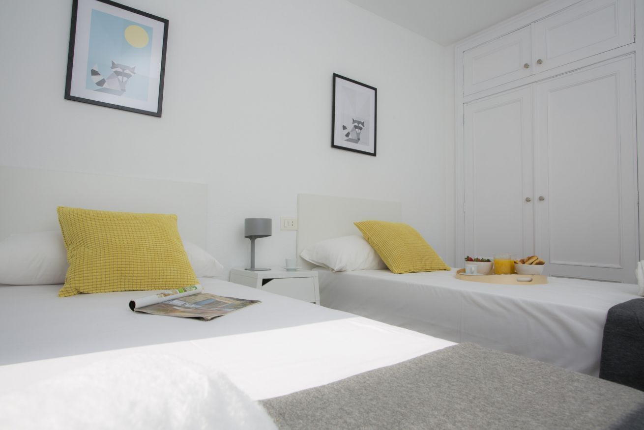 A hotel room with two beds, white bedding, and yellow pillows. There is a nightstand with a lamp and a tray with breakfast items.