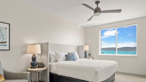 Ein Schlafzimmer mit Bett, Sessel, Nachttisch, Lampe, Deckenventilator und einem großen Fenster mit Meerblick.