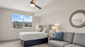 Ein Schlafzimmer mit einem Bett, einem grauen Sofa, einem runden Spiegel, einem Nachttisch mit Lampe und Blick auf Berge durch ein Fenster.