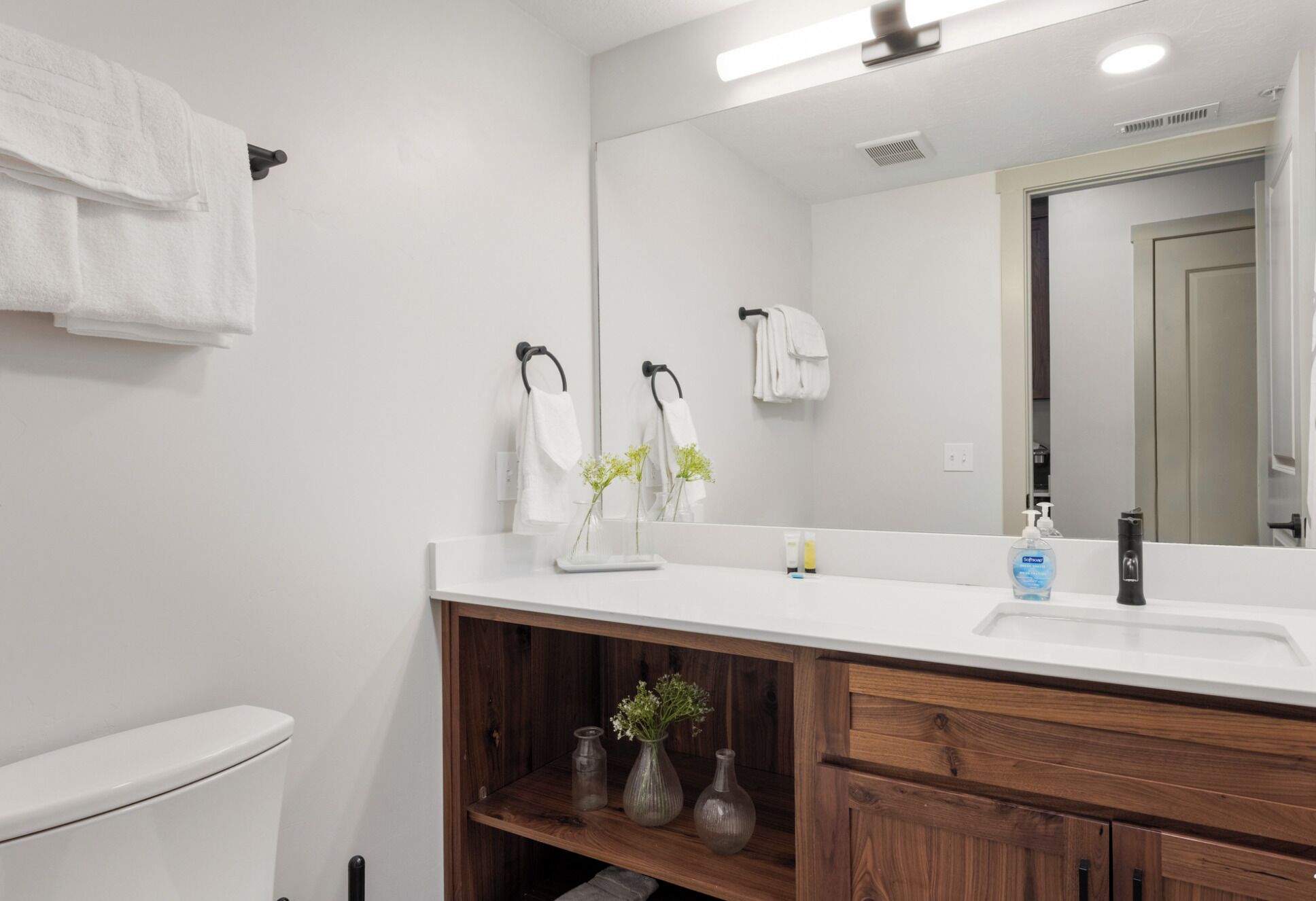 Une salle de bain avec des toilettes blanches, un meuble-lavabo en bois avec un miroir et une étagère ornée de vases.