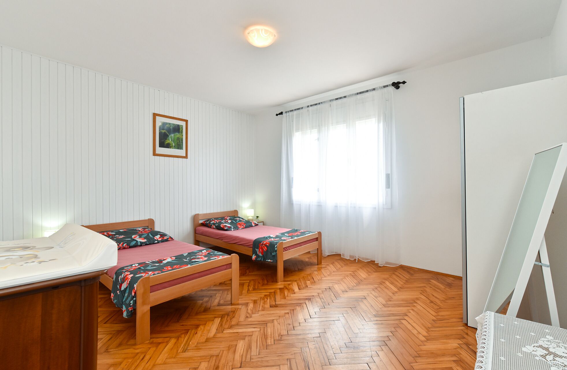 Quarto com duas camas de madeira, uma escrivaninha de madeira, um radiador branco e uma janela com cortinas translúcidas.