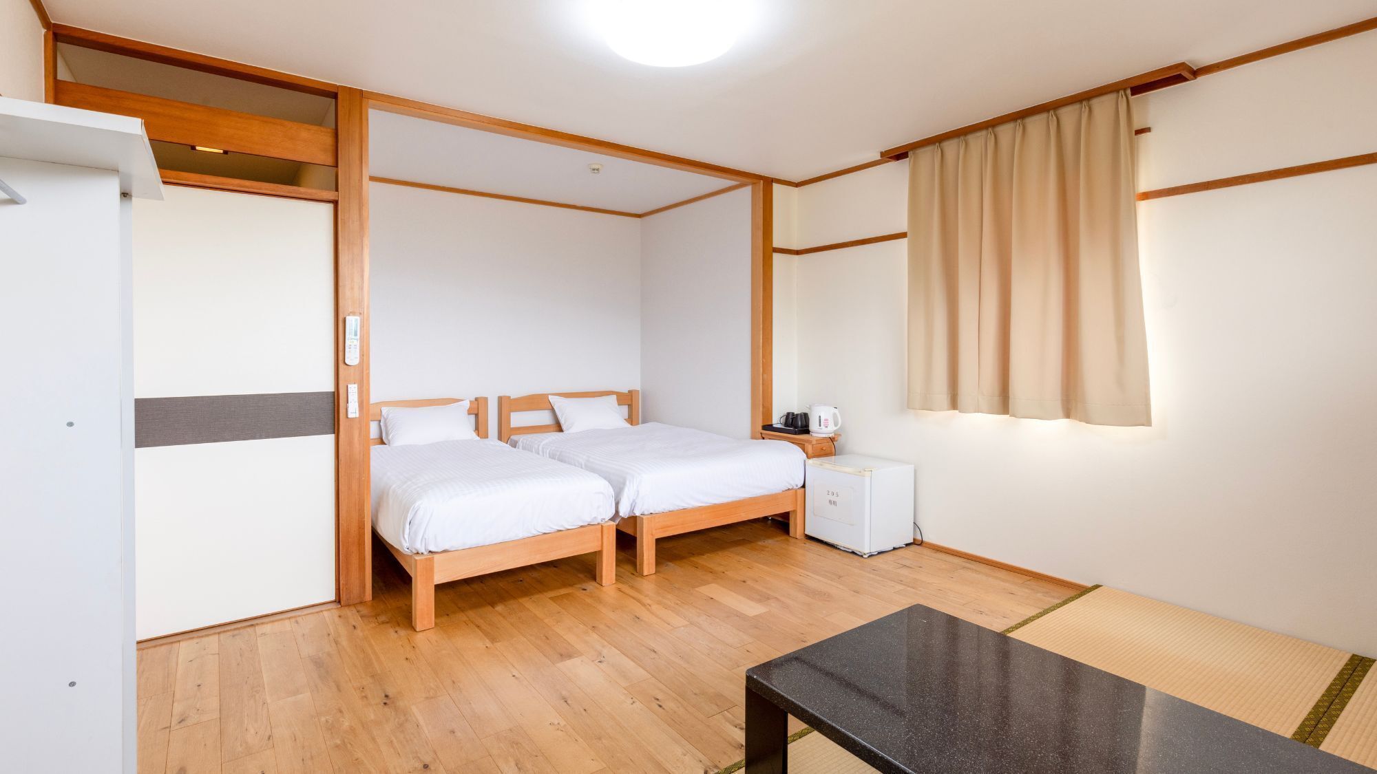 Una habitación con dos camas de madera, una mesita de noche con hervidor, un refrigerador y una ventana con cortinas.