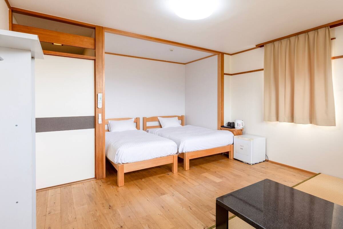 Una habitación con dos camas de madera, una mesita de noche con hervidor, un refrigerador y una ventana con cortinas.
