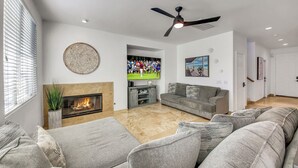 A modern living room with a fireplace, grey sofas, a flat-screen TV, and a ceiling fan.