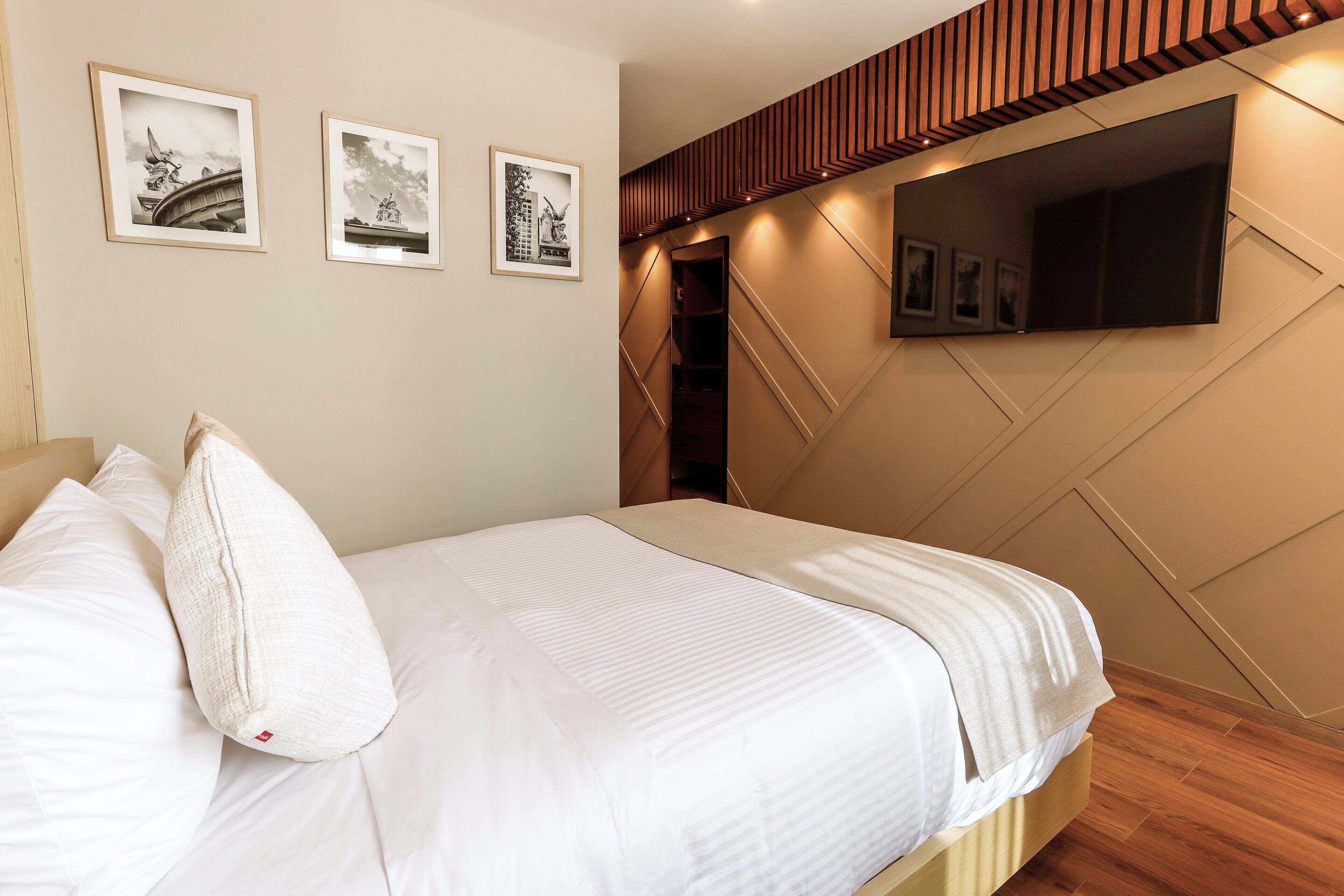 A neatly made bed with white linens and pillows, a wall-mounted TV, and framed pictures on the wall.