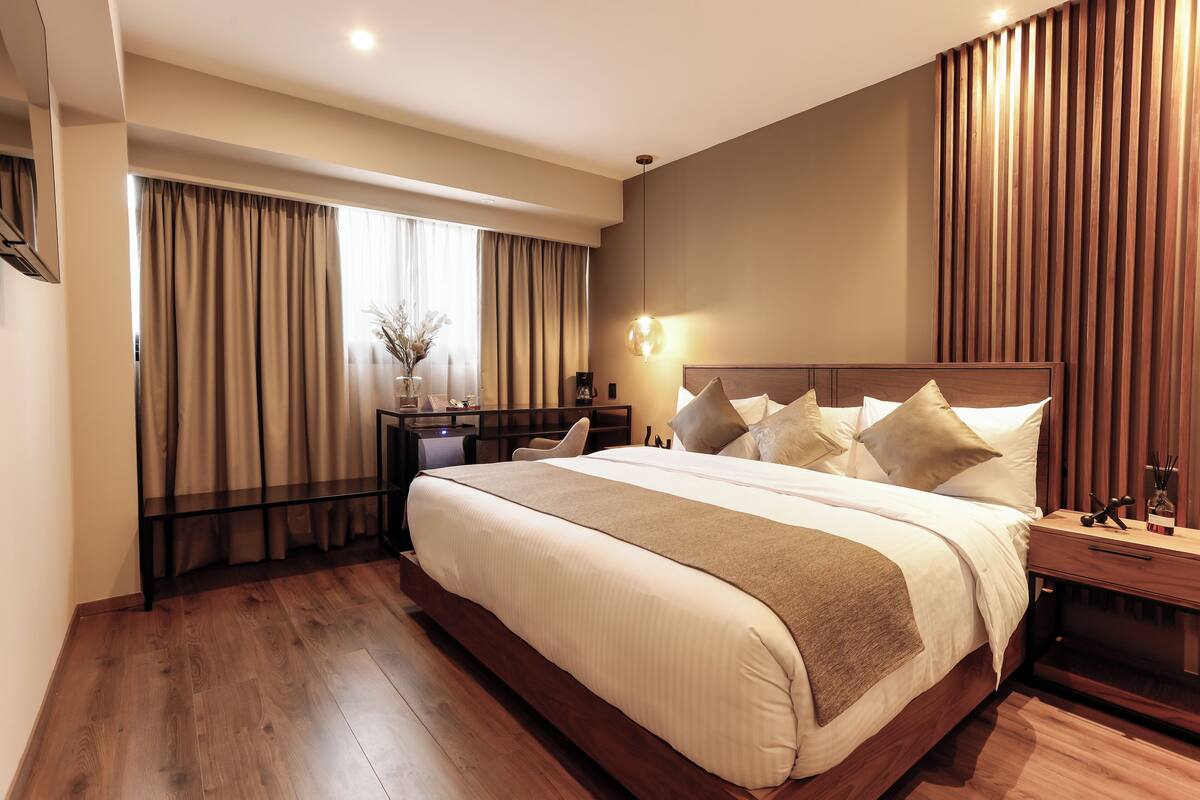 A modern hotel room with a large bed, wooden flooring, a bedside table with a lamp, and a view of the city through large windows.